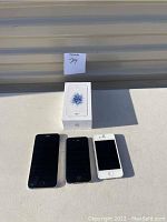 Three vintage iPhones displayed with a white iPhone SE box. iPhones include black iPhone 5, white iPhone 4S, and white iPhone SE (boxed).