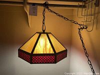 Photo of 1970s hanging ceiling lamp illuminated, showing amber glass panels and red glass section with diamond pattern in dark metal frame, hanging from chain with plug.