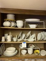 Full view of shelves showing multiple types of white porcelain dinnerware including stacked plates, cups hanging from a rack, ramekins, bowls, and platters.