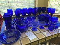 Wide view showing numerous blue cobalt glass goblets, pint glasses, bowls and cups on a tiled countertop next to window blinds.