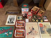 Photo showing a variety of Coca Cola collectibles including vintage magazine ads, coasters, tins, wooden boxes, signs for Budweiser and Billiards, and miscellaneous items.