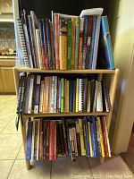 Front view of light wood bookshelf holding a thick assortment of cookbooks on three shelves