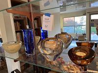 Four art glass candle holders including two Kosta Boda, one Orrefors, and one Waterford on glass shelving, showcasing shapes and colors.