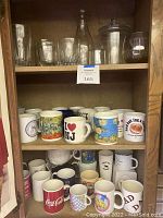 Shelf displaying 28+ novelty mugs featuring printed art and slogans such as Coca Cola logo, I Love NJ, Impressionist art, and Simpson's characters. Mugs vary in color and style, mostly ceramic.