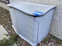 Grey Rubbermaid outdoor storage unit seen from the front showing closed unit against a wall outdoors.