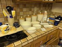 Wide view of kitchen counter showing assortment of ceramic kitchenware, canisters with wooden lids, cake molds, mugs, and utensils.
