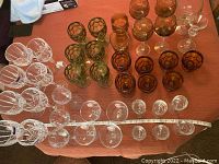 Amber and green colored assorted glasses including etched grape motif amber wine glasses, green textured all-purpose glasses, and some clear glasses on an orange cloth with tape measure for size reference.