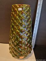 Textured ceramic vase with diamond/pine cone scale pattern in green and brown glaze with measuring tape showing height approximately 14 inches.