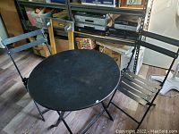 Round black metal bistro table with two matching black metal folding chairs placed around it on a wooden floor in a storage room.