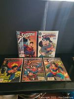 Five Superman comic books arranged in two rows on black surface, showing front covers. Titles include Superman: The Man of Steel and other Superman comics.