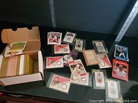 Photo showing a cardboard box filled with hundreds of baseball cards alongside a scattered selection of cards on a table, some in protective sleeves.