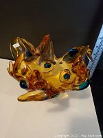 Front view showing the ruffled edges, blue oval accents and amber-colored glass body, with a ruler for scale