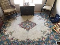 Full view of the rectangular area rug placed between two chairs and a small table, showing the central floral medallion and border design in cream, blue and pink hues.