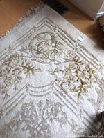 Close-up image showing off-white wool rug with gold floral and geometric raised design pattern, with visible fringe edge and some wear.