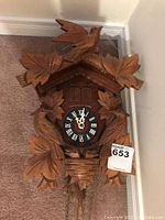 Front view of carved cuckoo clock laid on carpet floor, showing carved bird and leaf motifs around clock face with Roman numerals.