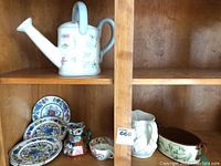 Wide view of top and bottom shelves showing multiple ceramic items including the Pfaltzgraff watering can on top left shelf and a grouping of plates, jugs and pot on the bottom right shelf.