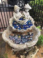 Front view of concrete two-tier outdoor fountain with two children figurines holding a bowl filled with blue and gold glass beads. Lower tier also filled with glass beads.