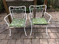 Front view of two white metal patio chairs with green floral cushions placed outdoors on a stone patio, showing the overall design and condition.