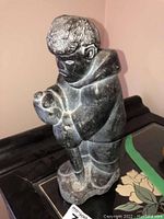 Front side view of soapstone carving showing standing Inuit figure with detailed hair and clothing, holding an object.