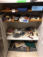 Metal cabinet shelves containing mixed hand tools including saws, circular saw blade, clamps, and sharpening stones.