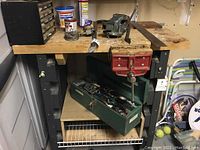 Wide view of workbench with mounted red bench vise and open green metal toolbox containing hand tools.