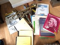 Trumpet placed on floor with stacked music books around it including Moonlight Sonata and Mozart volumes, and a small metronome.
