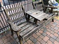 Full view of a weathered wood bench with built-in center table, slatted backrest and armrests, showing distressed condition of wood surface.