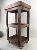 Front angled view showing entire wooden shelf unit with two shelves and top thick frame, on caster wheels