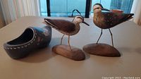 Photo showing two carved and painted wooden shore bird figurines and a painted wooden shoe on a flat surface.