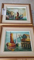 Both framed oil paintings stacked, showing Asian junks docked near wooden buildings and calm waters. Gold-tone wooden frames with white mats.