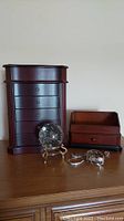 Front view of wooden five drawer jewelry box with side panels and matching smaller jewelry box with six pieces of silver-tone costume jewelry and glass ball on stand.