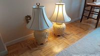 Two cream ceramic table lamps with fabric bell-shaped shades on wood floor near wall