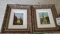 Two framed oil paintings side by side showing cityscape scenes under glass, both with carved ornate gold frames and white mats.