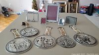 Full view of five pewter wall plaques with ceramic floral handles and four metal photo frames on a table.