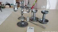 Photo showing set of pewter candle holders: a five-arm candelabra, two single candle holders (one with glass shade), and a candle snuffer on a beige table.