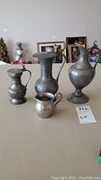 Four pewter items: 3 lidded jugs of varying sizes and 1 small creamer jug, all displayed on a beige surface with background items visible.