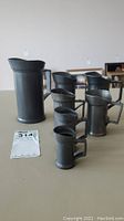 Front view showing all seven pewter measuring jugs of varying sizes aligned together with auction number card in front.