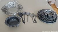 Overview of all pewter pieces showing pedestal bowl, footed bowl, stack of plates, and serving spoons