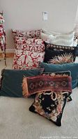 Group of various decorative pillows in different patterns and colors including damask print, floral embroidery, solid teal, and textured cream fabric with two smaller pillows with fringe