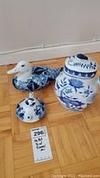 Photo of a blue and white decorated duck-shaped ceramic teapot, a lidded jar with blue floral patterns, and a small teapot lid on the floor.