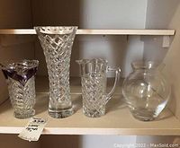 Four assorted clear glass vases of various shapes and sizes on shelf.