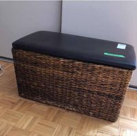 Frontal view of the wicker storage chest showing the woven construction and the padded black seat top.