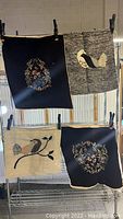 Four handmade wall hangings hanging on a rack. Two with floral embroidery on dark fabric; one with bird on gray; one with bird on beige.