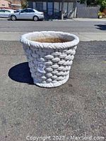White plaster pot with woven basket pattern and rope rim, with visible weathering and erosion on base, placed on asphalt outdoors