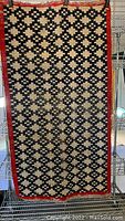 Full view of the rectangular area rug showing the black geometric pattern on beige with a red border, standing vertically on a metal rack.
