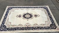 Full view of rectangular cream and blue floral patterned area rug laid out on pavement, showing central medallion and border.
