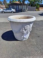Full side view of large white plaster decorative pot showing scalloped rim and embossed grape cluster and floral designs.