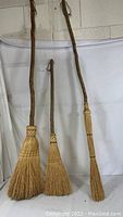 Three handmade brooms with natural wood branch handles and straw bristles, arranged against a white backdrop.