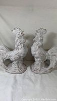 Front view of two ceramic rooster statues, white with distressed detailing, standing side by side on a white cloth background.