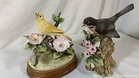 Front view of both bird figurines placed side by side, showing the Robin on a tree stump and Canary on a branch with flowers on wooden base.